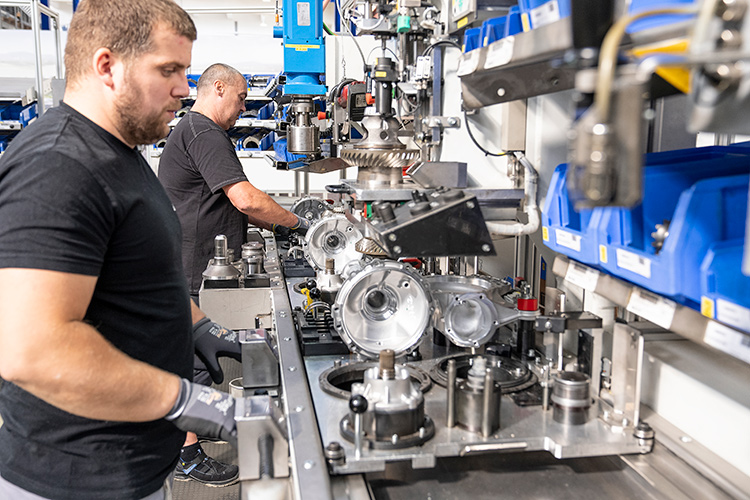 Two factory workers operating machinery to assemble metal automotive components.