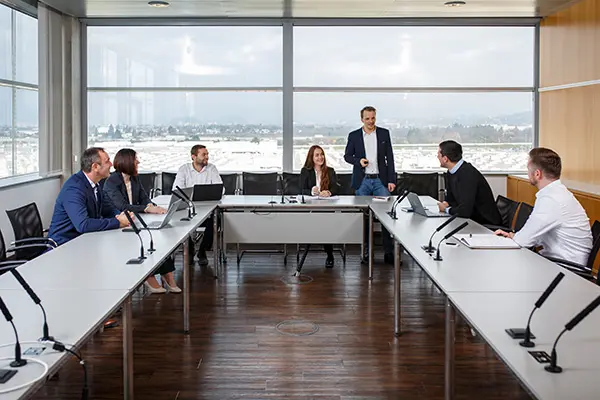 Business meeting with seven people seated around a conference table, one person standing and presenting