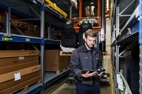 Worker using a tablet in a warehouse aisle with car seats and boxes on shelves